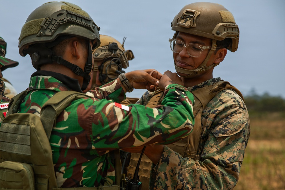 Two military personnel from different nations are engaged in what appears to be a pin or patch exchange ceremony, a common gesture of goodwill during joint military exercises. The soldier on the left wears Indonesian Army (TNI-AD) camouflage with a visible 'INDONESIA' name tape and red-and-white flag patch. The soldier on the right wears US Marine Corps MARPAT digital camouflage with a tan tactical vest and ballistic helmet.