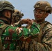 Two military personnel from different nations are engaged in what appears to be a pin or patch exchange ceremony, a common gesture of goodwill during joint military exercises. The soldier on the left wears Indonesian Army (TNI-AD) camouflage with a visible 'INDONESIA' name tape and red-and-white flag patch. The soldier on the right wears US Marine Corps MARPAT digital camouflage with a tan tactical vest and ballistic helmet.
