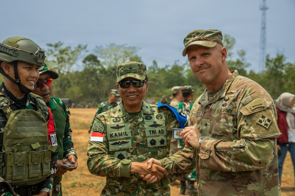 Two military officers — one from the Indonesian Army (TNI-AD) and one from the U.S. Army — are shaking hands in what appears to be a joint military exercise or ceremony. The Indonesian officer, identified by his name tape as 'KAMIL' and wearing a TNI-AD patch with an Indonesian flag, is exchanging what appears to be a challenge coin or medallion with the U.S. Army officer. Additional military personnel are visible in the background.