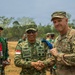 Two military officers — one from the Indonesian Army (TNI-AD) and one from the U.S. Army — are shaking hands in what appears to be a joint military exercise or ceremony. The Indonesian officer, identified by his name tape as 'KAMIL' and wearing a TNI-AD patch with an Indonesian flag, is exchanging what appears to be a challenge coin or medallion with the U.S. Army officer. Additional military personnel are visible in the background.