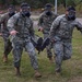 Four military personnel wearing MARPAT digital camouflage uniforms and full-face gas masks are conducting a casualty evacuation drill, carrying an incapacitated person across a grassy field. The exercise appears to be a CBRN (Chemical, Biological, Radiological, Nuclear) training scenario based on the protective masks worn. Additional personnel in different camouflage uniforms are visible in the background, suggesting a multinational training exercise.