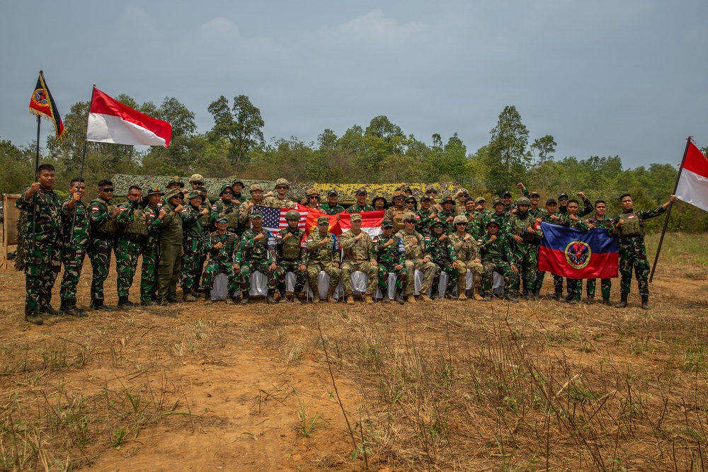 A large group of military personnel from multiple nations pose together for a group photograph at an outdoor training or exercise location. The group includes soldiers in Indonesian TNI camouflage uniforms, US military personnel in MultiCam/OCP uniforms, and others, gathered in front of camouflage-netted military vehicles or structures. Multiple national flags are visible, indicating a multinational military exercise or joint training event.