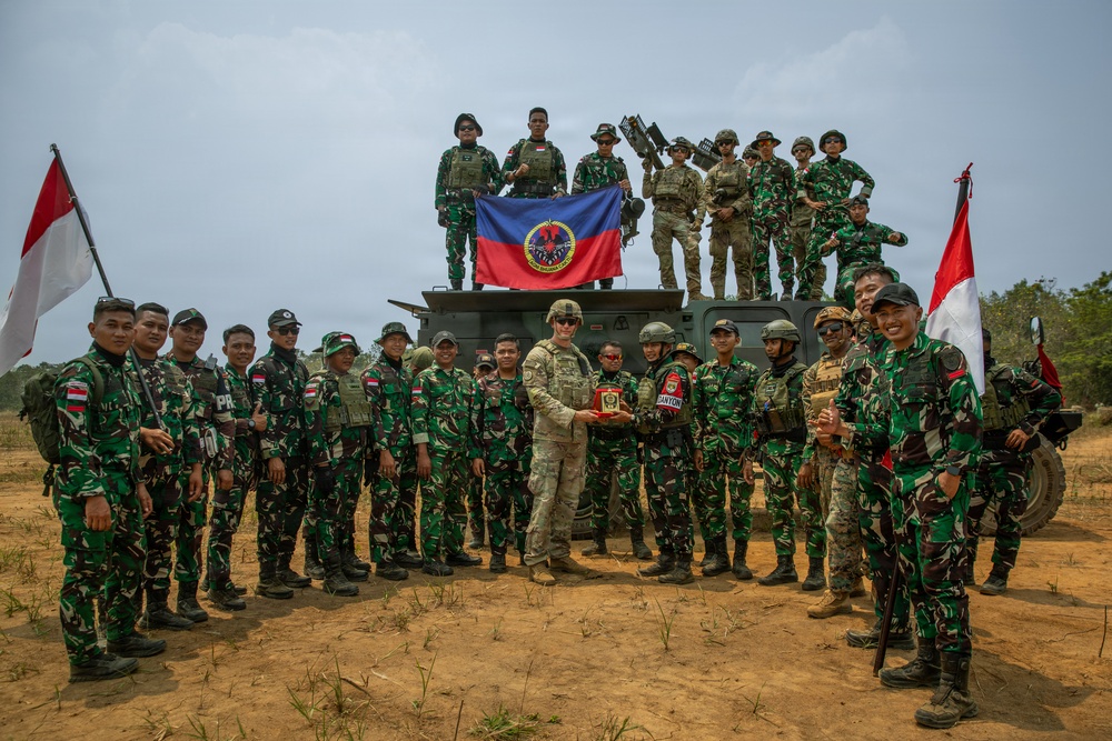 A group photo of military personnel from what appears to be a joint exercise between Indonesian Armed Forces (TNI) and US military personnel. Soldiers are posed in front of and on top of a military vehicle, with Indonesian flags and a unit banner visible. Two soldiers in the center appear to be exchanging a commemorative plaque or gift.