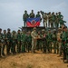 A group photo of military personnel from what appears to be a joint exercise between Indonesian Armed Forces (TNI) and US military personnel. Soldiers are posed in front of and on top of a military vehicle, with Indonesian flags and a unit banner visible. Two soldiers in the center appear to be exchanging a commemorative plaque or gift.