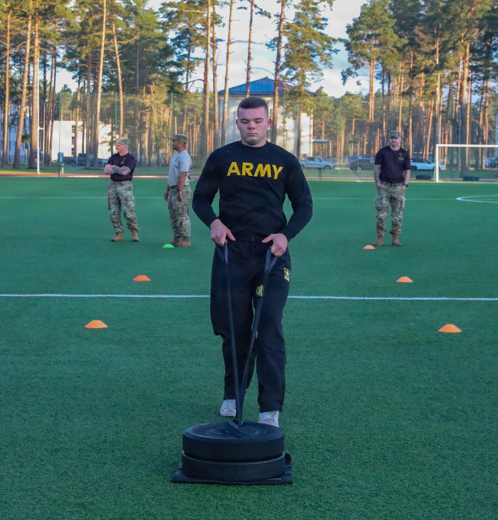 A U.S. Army soldier in black PT (physical training) uniform is performing a sled drag exercise on an artificial turf field, pulling straps attached to a weighted sled. Several observers in military uniforms and civilian attire stand in the background watching. The setting appears to be a sports facility surrounded by tall pine trees.