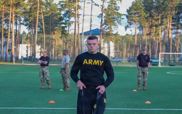 A U.S. Army soldier in black PT (physical training) uniform is performing a sled drag exercise on an artificial turf field, pulling straps attached to a weighted sled. Several observers in military uniforms and civilian attire stand in the background watching. The setting appears to be a sports facility surrounded by tall pine trees.