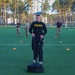 A U.S. Army soldier in black PT (physical training) uniform is performing a sled drag exercise on an artificial turf field, pulling straps attached to a weighted sled. Several observers in military uniforms and civilian attire stand in the background watching. The setting appears to be a sports facility surrounded by tall pine trees.