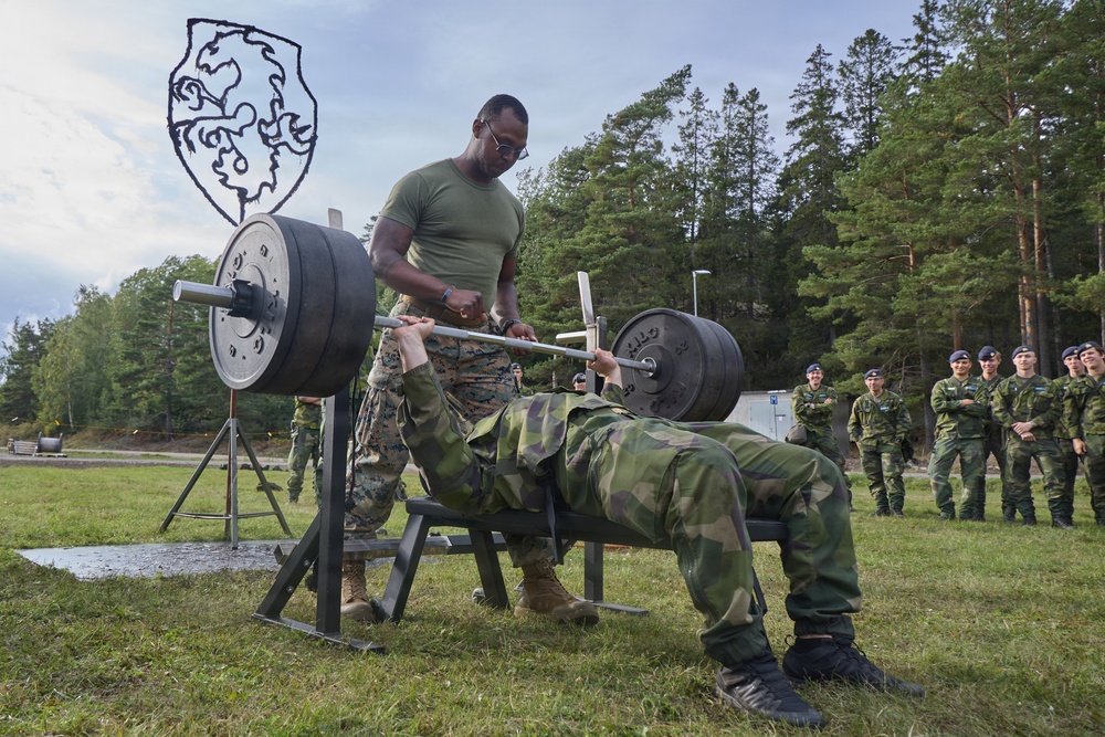 A military physical fitness exercise is taking place outdoors, where a soldier in camouflage uniform performs a bench press while being spotted by another soldier in a green t-shirt and camouflage pants. A group of uniformed military personnel observes the activity in the background. The setting appears to be a military training area surrounded by pine trees, with a shield-shaped emblem visible in the background.