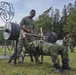 A military physical fitness exercise is taking place outdoors, where a soldier in camouflage uniform performs a bench press while being spotted by another soldier in a green t-shirt and camouflage pants. A group of uniformed military personnel observes the activity in the background. The setting appears to be a military training area surrounded by pine trees, with a shield-shaped emblem visible in the background.