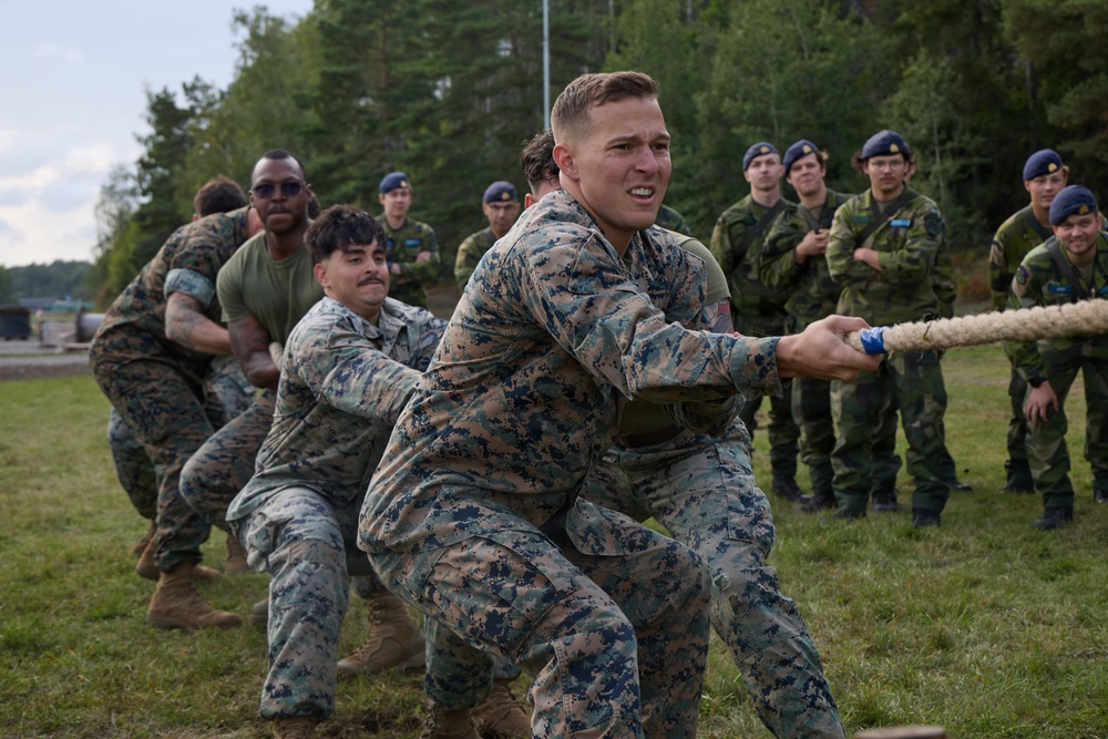 A group of military personnel in US Marine Corps digital camouflage uniforms are competing in a tug-of-war against what appears to be Swedish military personnel wearing woodland camouflage and dark blue berets. The event appears to be a friendly inter-military competition or joint exercise, likely held at a training facility in Scandinavia. Spectators in Swedish military uniforms observe from the sidelines.