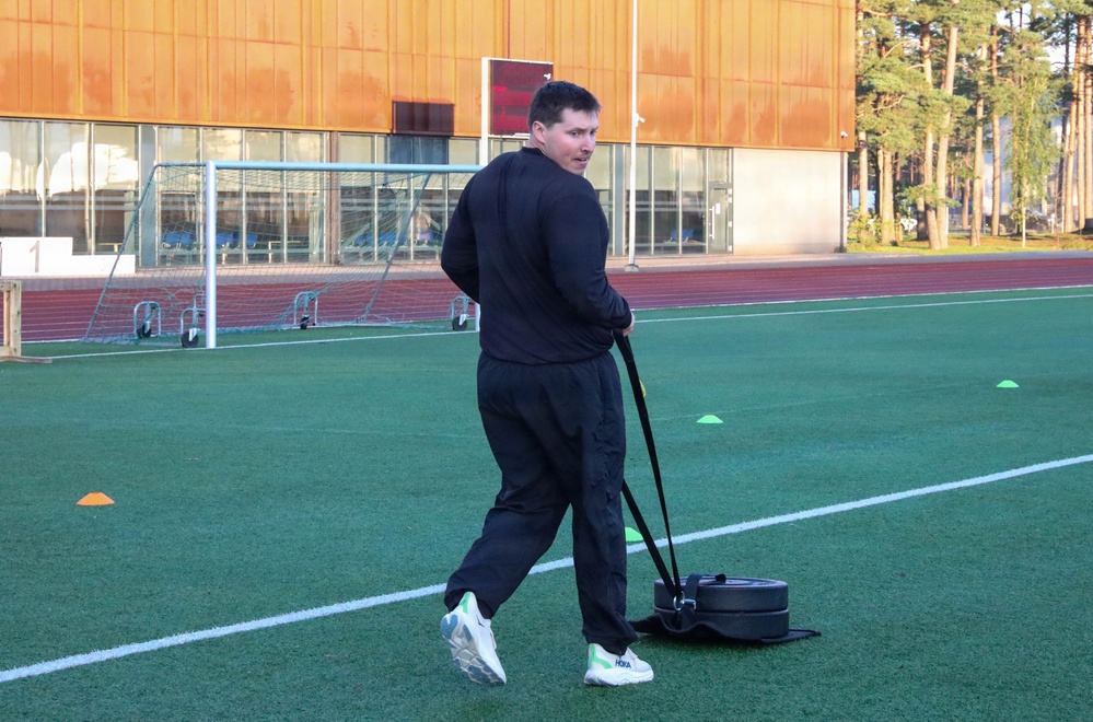 A man dressed in a black tracksuit is performing a sled drag exercise on an artificial turf sports field. He is pulling a weighted sled using straps while looking back over his shoulder. The setting appears to be a multi-use athletic facility with a running track, soccer goals, and a modern building in the background.