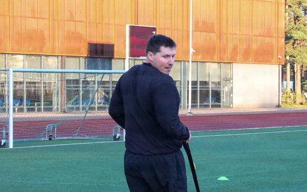 A man dressed in a black tracksuit is performing a sled drag exercise on an artificial turf sports field. He is pulling a weighted sled using straps while looking back over his shoulder. The setting appears to be a multi-use athletic facility with a running track, soccer goals, and a modern building in the background.