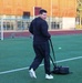 A man dressed in a black tracksuit is performing a sled drag exercise on an artificial turf sports field. He is pulling a weighted sled using straps while looking back over his shoulder. The setting appears to be a multi-use athletic facility with a running track, soccer goals, and a modern building in the background.