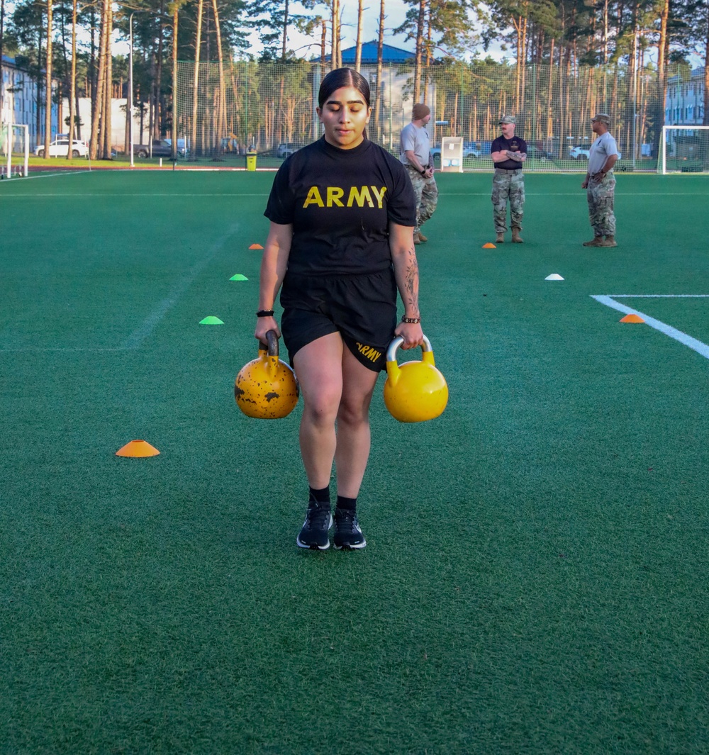 A U.S. Army soldier in standard PT (Physical Training) uniform is performing a kettlebell carry exercise on an artificial turf field, holding one kettlebell in each hand. The exercise appears to be part of a formal fitness assessment or training event, with several uniformed observers visible in the background. Colored cone markers are placed on the field to designate the course or exercise boundaries.