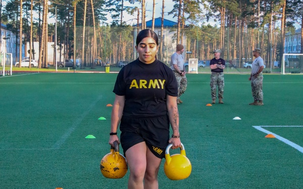 A U.S. Army soldier in standard PT (Physical Training) uniform is performing a kettlebell carry exercise on an artificial turf field, holding one kettlebell in each hand. The exercise appears to be part of a formal fitness assessment or training event, with several uniformed observers visible in the background. Colored cone markers are placed on the field to designate the course or exercise boundaries.