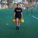 A U.S. Army soldier in standard PT (Physical Training) uniform is performing a kettlebell carry exercise on an artificial turf field, holding one kettlebell in each hand. The exercise appears to be part of a formal fitness assessment or training event, with several uniformed observers visible in the background. Colored cone markers are placed on the field to designate the course or exercise boundaries.
