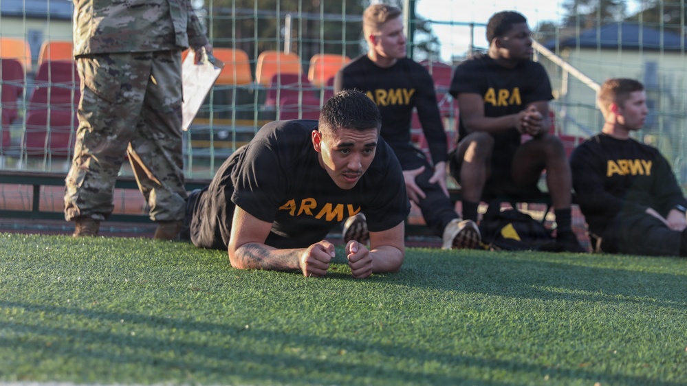 A group of U.S. Army soldiers in black PT (physical training) uniforms are on an artificial turf field, likely conducting a fitness test or physical training session. One soldier in the foreground is performing a plank exercise while others rest near a fence with stadium seating visible in the background. A soldier in camouflage uniform, presumably an evaluator or instructor, is visible in the upper left holding papers.