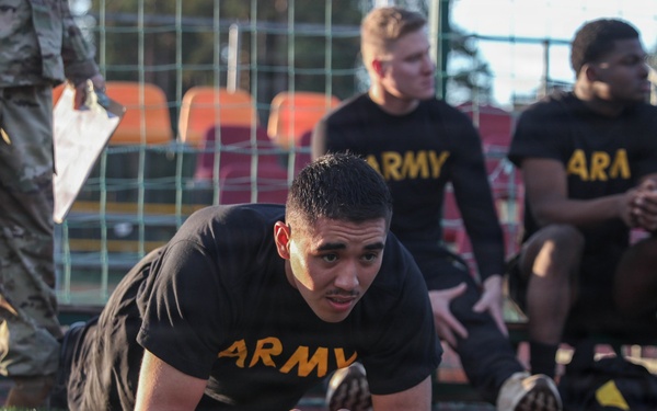 A group of U.S. Army soldiers in black PT (physical training) uniforms are on an artificial turf field, likely conducting a fitness test or physical training session. One soldier in the foreground is performing a plank exercise while others rest near a fence with stadium seating visible in the background. A soldier in camouflage uniform, presumably an evaluator or instructor, is visible in the upper left holding papers.