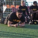 A group of U.S. Army soldiers in black PT (physical training) uniforms are on an artificial turf field, likely conducting a fitness test or physical training session. One soldier in the foreground is performing a plank exercise while others rest near a fence with stadium seating visible in the background. A soldier in camouflage uniform, presumably an evaluator or instructor, is visible in the upper left holding papers.