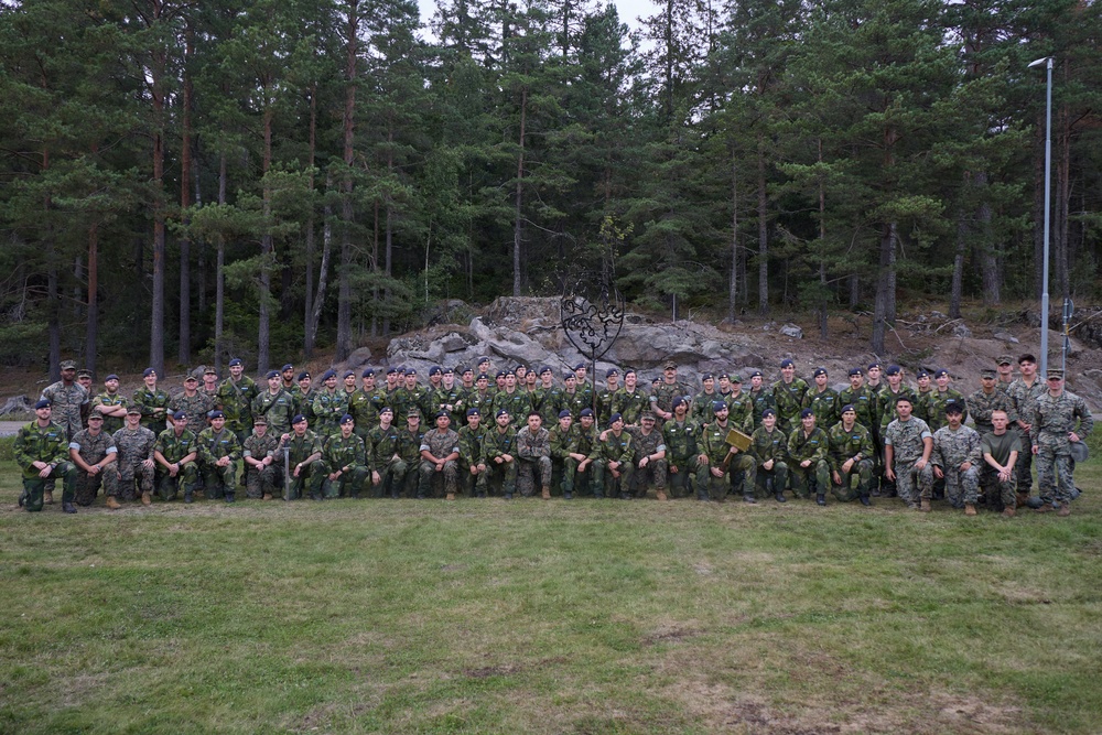 A large group of military personnel from multiple nations poses for a formal group photograph outdoors. The soldiers are wearing a variety of camouflage uniforms and berets, suggesting a multinational military exercise or joint training event. They are arranged in two rows — standing and kneeling — in front of a rocky outcrop and pine forest backdrop.