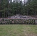 A large group of military personnel from multiple nations poses for a formal group photograph outdoors. The soldiers are wearing a variety of camouflage uniforms and berets, suggesting a multinational military exercise or joint training event. They are arranged in two rows — standing and kneeling — in front of a rocky outcrop and pine forest backdrop.