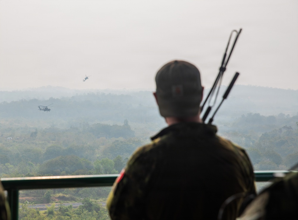 A soldier in military fatigues stands on an elevated observation platform, watching two helicopters flying over a hazy, forested landscape. The soldier is viewed from behind and is carrying what appears to be multiple rifle barrels or antenna equipment on their back. The scene suggests a military observation or patrol activity in a tropical or subtropical region.