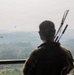 A soldier in military fatigues stands on an elevated observation platform, watching two helicopters flying over a hazy, forested landscape. The soldier is viewed from behind and is carrying what appears to be multiple rifle barrels or antenna equipment on their back. The scene suggests a military observation or patrol activity in a tropical or subtropical region.