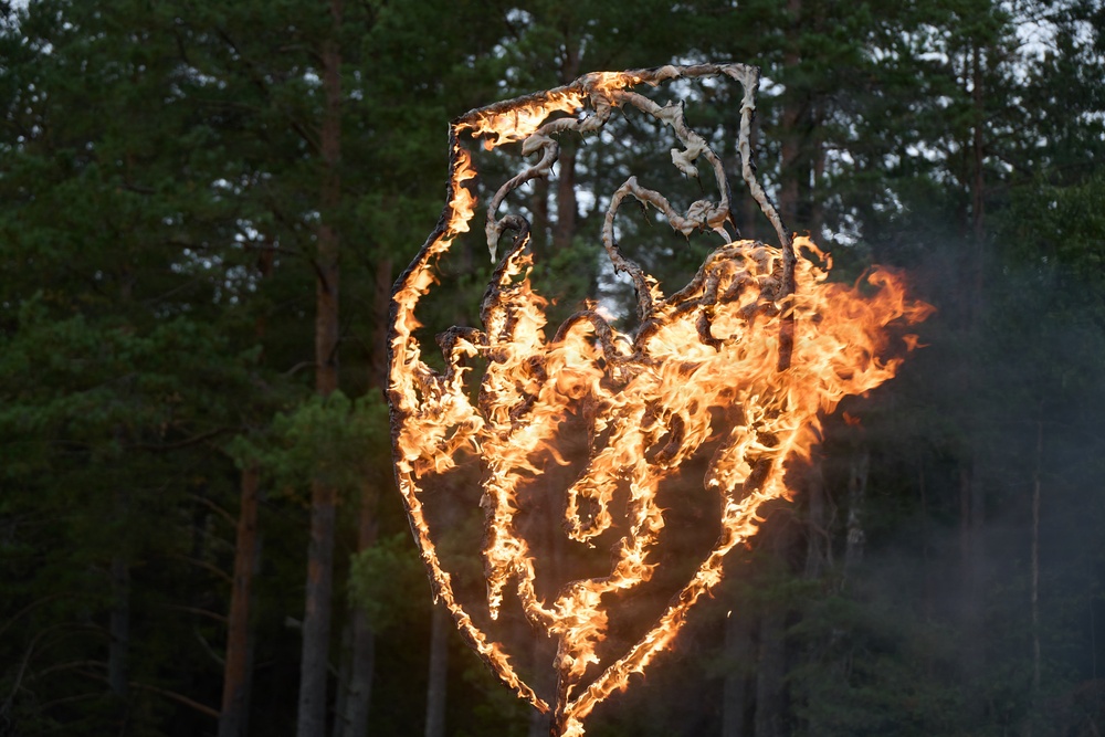 A large sculptural or decorative structure constructed from woven or twisted organic material (likely birch bark or similar) is actively burning with bright orange flames. The structure appears to be heart-shaped or shield-shaped with intricate internal patterns, possibly resembling a face or symbolic design. It is set against a backdrop of dense coniferous forest in dim, overcast lighting.