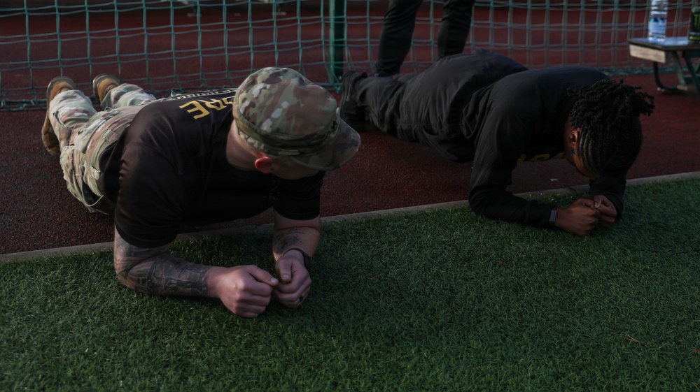 Two individuals are performing plank exercises on an artificial turf surface near a track and field area. The person on the left is wearing a military-style multicam cap, a dark t-shirt, and combat boots, with visible tattoos on their forearms. Both are in a forearm plank position, suggesting a physical fitness training session.
