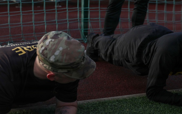 Two individuals are performing plank exercises on an artificial turf surface near a track and field area. The person on the left is wearing a military-style multicam cap, a dark t-shirt, and combat boots, with visible tattoos on their forearms. Both are in a forearm plank position, suggesting a physical fitness training session.