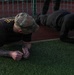 Two individuals are performing plank exercises on an artificial turf surface near a track and field area. The person on the left is wearing a military-style multicam cap, a dark t-shirt, and combat boots, with visible tattoos on their forearms. Both are in a forearm plank position, suggesting a physical fitness training session.