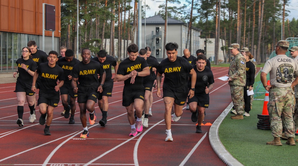 A group of U.S. Army personnel in black PT (Physical Training) uniforms are running on a red athletic track. On the right side of the track, uniformed cadre members in camouflage uniforms are observing and monitoring the run. The setting appears to be a military installation with buildings and tall pine trees visible in the background.