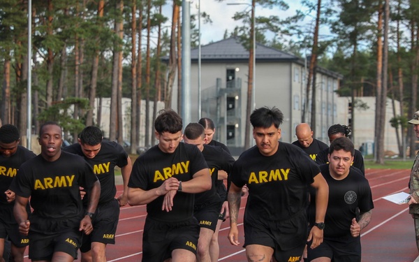A group of U.S. Army personnel in black PT (Physical Training) uniforms are running on a red athletic track. On the right side of the track, uniformed cadre members in camouflage uniforms are observing and monitoring the run. The setting appears to be a military installation with buildings and tall pine trees visible in the background.