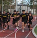 A group of U.S. Army personnel in black PT (Physical Training) uniforms are running on a red athletic track. On the right side of the track, uniformed cadre members in camouflage uniforms are observing and monitoring the run. The setting appears to be a military installation with buildings and tall pine trees visible in the background.