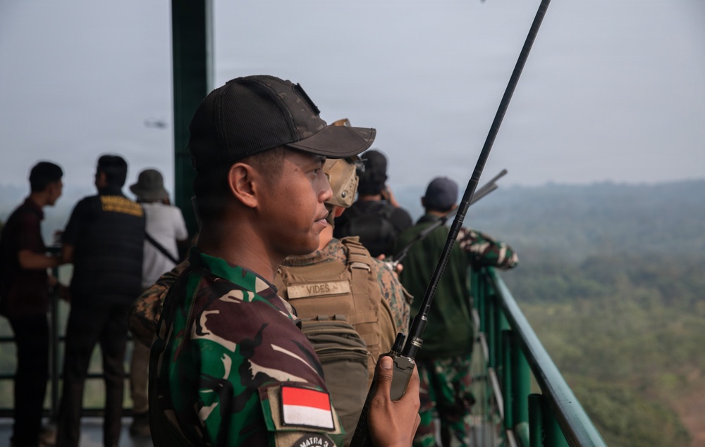 The image shows military personnel on an elevated observation platform overlooking a forested landscape. A soldier in the foreground wears Indonesian Army (TNI) camouflage with an Indonesian flag patch and holds a radio transceiver, while another individual behind him wears tan tactical gear with a name tape reading 'VIDES'. Multiple personnel are visible looking out over the terrain, suggesting a joint observation or coordination activity.