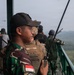 The image shows military personnel on an elevated observation platform overlooking a forested landscape. A soldier in the foreground wears Indonesian Army (TNI) camouflage with an Indonesian flag patch and holds a radio transceiver, while another individual behind him wears tan tactical gear with a name tape reading 'VIDES'. Multiple personnel are visible looking out over the terrain, suggesting a joint observation or coordination activity.