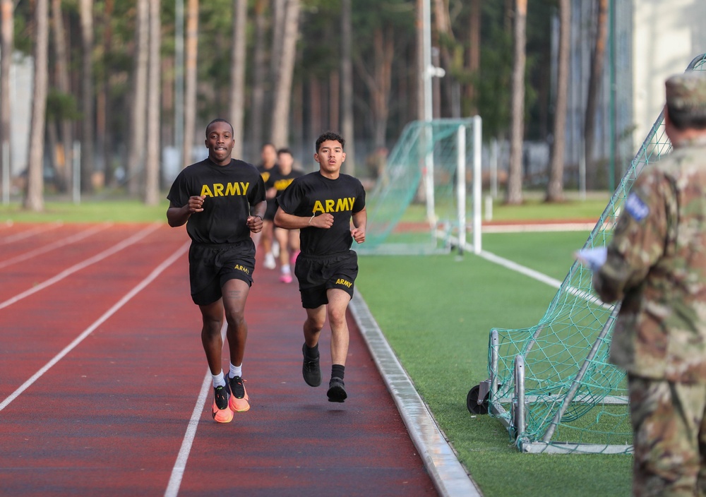 Several individuals wearing U.S. Army physical training uniforms (black shirts and shorts with yellow 'ARMY' lettering) are running on a red athletic track. Two runners are prominently in the foreground, with additional runners visible behind them. A person in military camouflage uniform stands to the right side, appearing to observe or monitor the run.