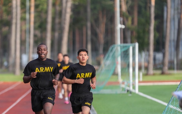 Several individuals wearing U.S. Army physical training uniforms (black shirts and shorts with yellow 'ARMY' lettering) are running on a red athletic track. Two runners are prominently in the foreground, with additional runners visible behind them. A person in military camouflage uniform stands to the right side, appearing to observe or monitor the run.