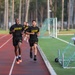 Several individuals wearing U.S. Army physical training uniforms (black shirts and shorts with yellow 'ARMY' lettering) are running on a red athletic track. Two runners are prominently in the foreground, with additional runners visible behind them. A person in military camouflage uniform stands to the right side, appearing to observe or monitor the run.