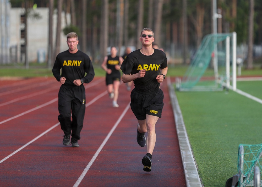 Multiple U.S. Army personnel are running on a red athletic track, wearing standard-issue black Army physical training uniforms with yellow lettering. The foreground runner is in shorts and a t-shirt with sunglasses, while others behind wear long pants and long-sleeve shirts. The setting appears to be a military base or installation with a sports field and trees visible in the background.