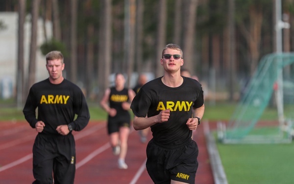 Multiple U.S. Army personnel are running on a red athletic track, wearing standard-issue black Army physical training uniforms with yellow lettering. The foreground runner is in shorts and a t-shirt with sunglasses, while others behind wear long pants and long-sleeve shirts. The setting appears to be a military base or installation with a sports field and trees visible in the background.