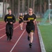 Multiple U.S. Army personnel are running on a red athletic track, wearing standard-issue black Army physical training uniforms with yellow lettering. The foreground runner is in shorts and a t-shirt with sunglasses, while others behind wear long pants and long-sleeve shirts. The setting appears to be a military base or installation with a sports field and trees visible in the background.