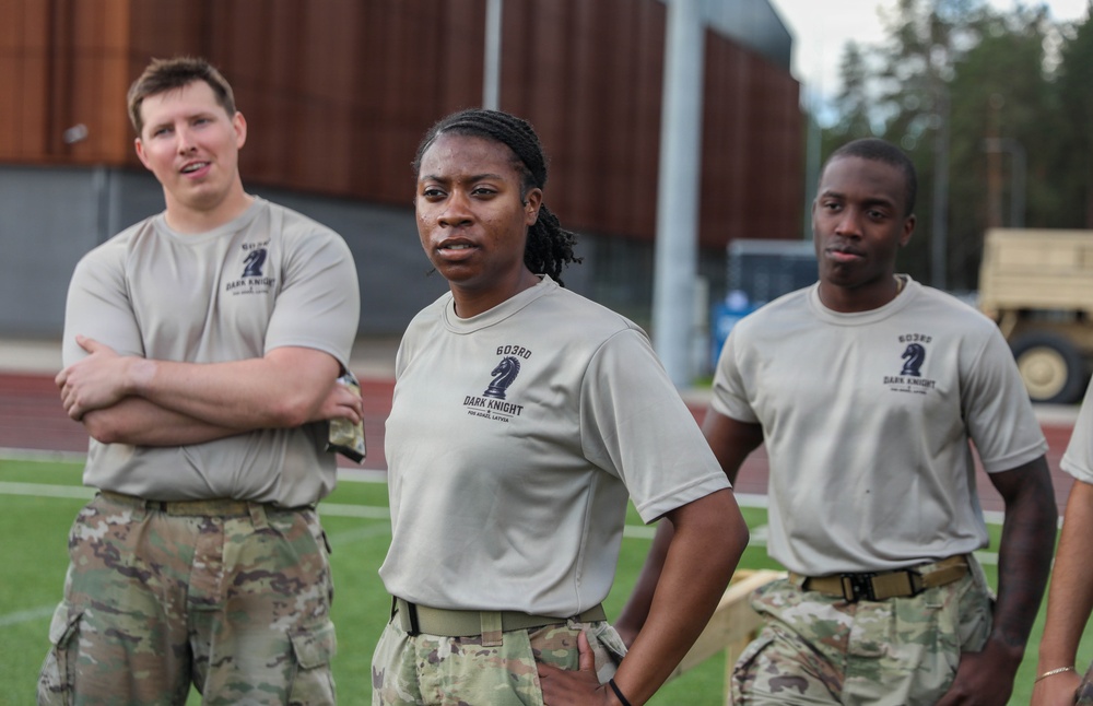 Three military personnel are standing on what appears to be a sports field or track, all wearing matching tan 'Dark Knight' unit t-shirts and camouflage (OCP/MultiCam) uniform bottoms. The shirts display '603RD DARK KNIGHT' text with a chess knight piece logo. They appear to be at an outdoor physical training or unit event.