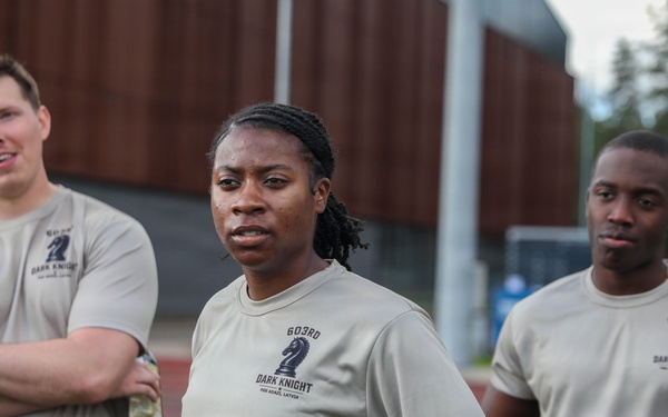 Three military personnel are standing on what appears to be a sports field or track, all wearing matching tan 'Dark Knight' unit t-shirts and camouflage (OCP/MultiCam) uniform bottoms. The shirts display '603RD DARK KNIGHT' text with a chess knight piece logo. They appear to be at an outdoor physical training or unit event.