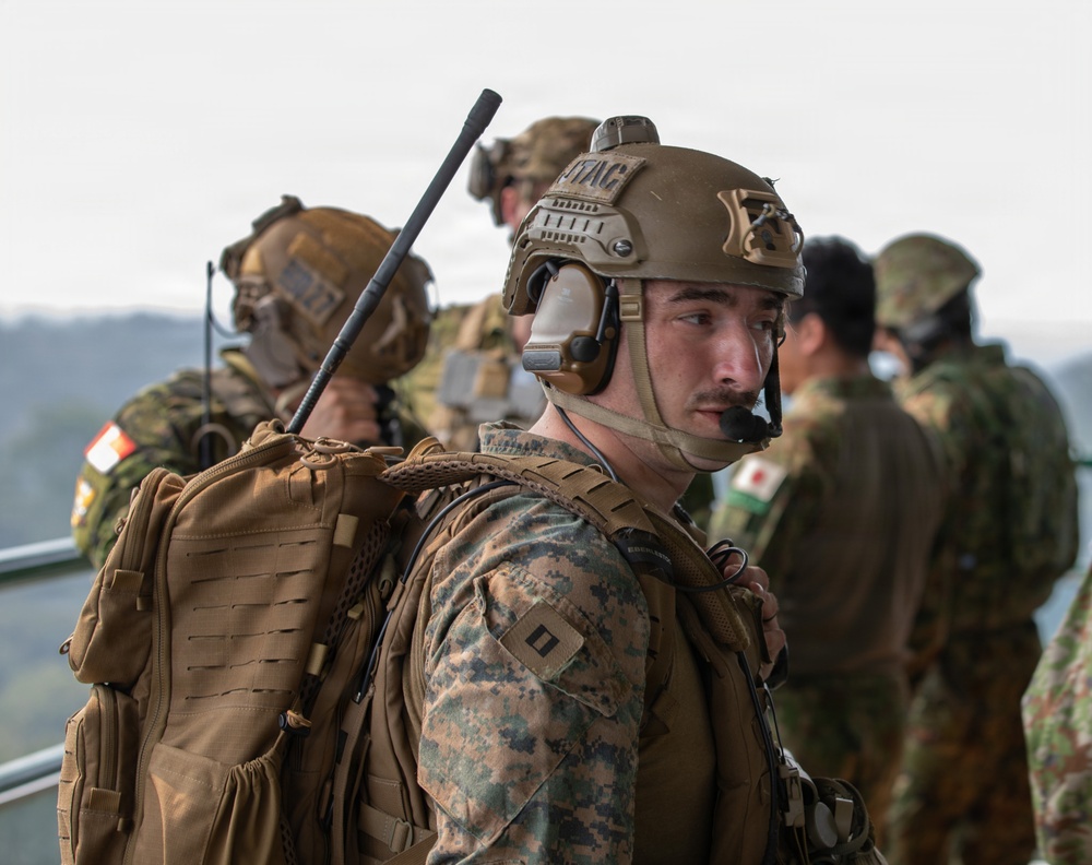 The image shows military personnel from what appears to be a joint U.S. Marine Corps and Japanese Ground Self-Defense Force exercise or operation. The foreground features a U.S. Marine wearing MARPAT camouflage, a tan tactical helmet with 'JTAC' label, communications headset, and a tactical backpack with a radio antenna. In the background, personnel in Japanese military uniforms with Japanese flag patches are visible alongside other U.S. Marines.