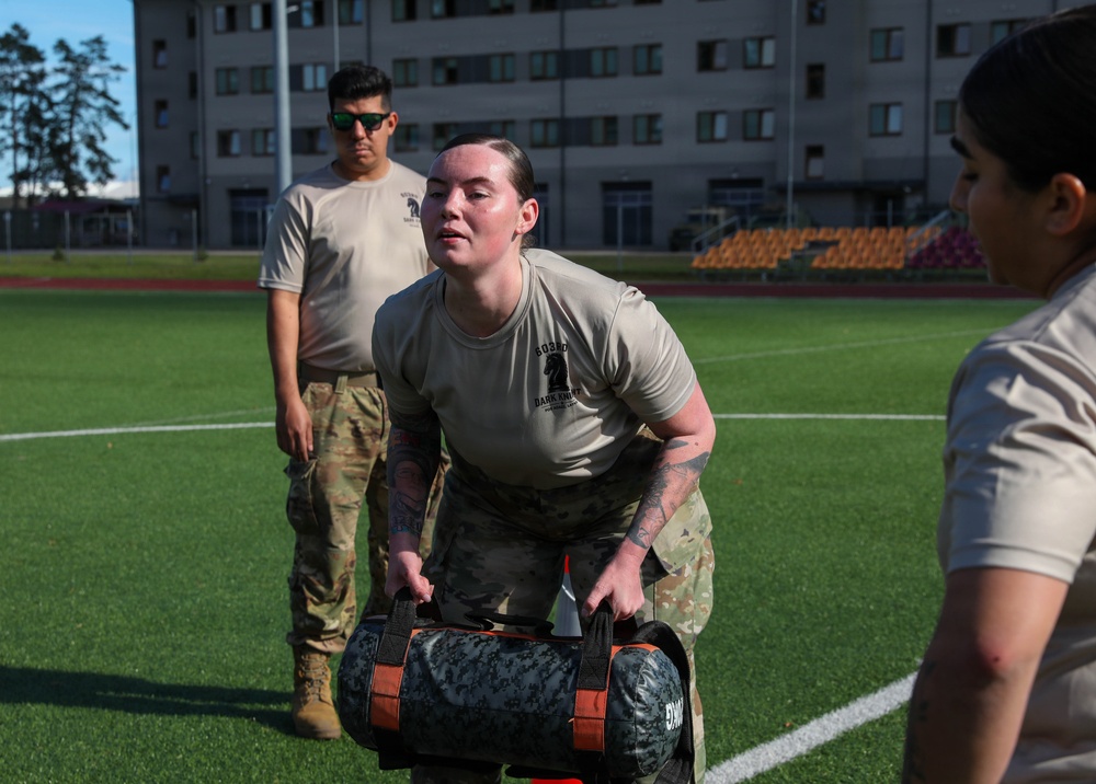 Military personnel are conducting a physical fitness training exercise on an outdoor athletic field. A female soldier in the foreground is lifting or carrying a heavy weighted duffel/sandbag, while two other soldiers observe in the background. The setting appears to be a military installation with a stadium or sports facility visible in the background.
