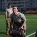 Military personnel are conducting a physical fitness training exercise on an outdoor athletic field. A female soldier in the foreground is lifting or carrying a heavy weighted duffel/sandbag, while two other soldiers observe in the background. The setting appears to be a military installation with a stadium or sports facility visible in the background.