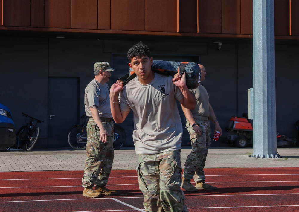 A military service member in camouflage pants and a light gray t-shirt carries a weighted sandbag across his shoulders on a red running track. Two other uniformed personnel in full camouflage uniforms stand in the background near a building. The scene appears to be a physical fitness or training exercise at a military installation.