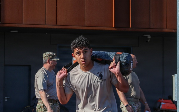 A military service member in camouflage pants and a light gray t-shirt carries a weighted sandbag across his shoulders on a red running track. Two other uniformed personnel in full camouflage uniforms stand in the background near a building. The scene appears to be a physical fitness or training exercise at a military installation.