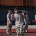 A military service member in camouflage pants and a light gray t-shirt carries a weighted sandbag across his shoulders on a red running track. Two other uniformed personnel in full camouflage uniforms stand in the background near a building. The scene appears to be a physical fitness or training exercise at a military installation.